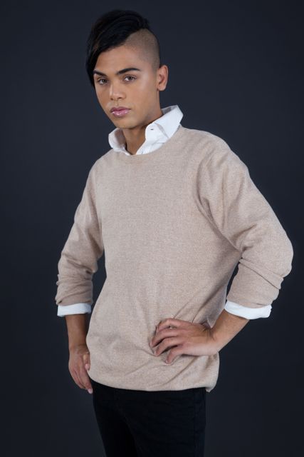 Androgynous person posing confidently with hands on hips against a grey background. Wearing a beige sweater and black pants, this image highlights modern fashion and unique style. Ideal for use in fashion blogs, gender-neutral clothing advertisements, and articles on androgyny and self-expression.