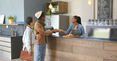 Young diverse couple paying friendly barista while receiving pastry at cozy urban cafe counter