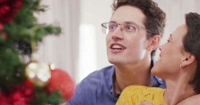 Couple wearing casual clothing and smiling while decorating a Christmas tree with ornaments. Ideal for holiday cards, festive promotions, and family-centric advertising celebrating warmth and togetherness.