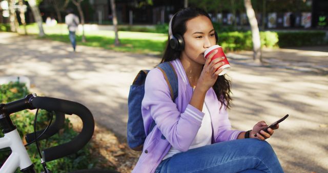 Young woman is drinking coffee and listening to music in a park setting. She wears headphones and has a backpack, suggesting she might be a student or someone enjoying a leisurely break. Ideal for concepts related to lifestyle, relaxation, outdoor activities, student life, and morning routines.