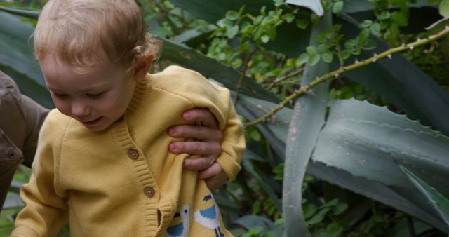 Capturing a tender moment, this image shows a parent holding their small child among large, lush greenery with tall plants in the background. Suitable for use in parenting blogs, family-related articles, nature and outdoors branding, and advertisements promoting children's clothing or family activities.