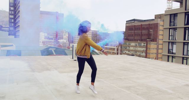 Young biracial woman celebrates outdoors with a smoke bomb. Her joyous moment is captured on a rooftop with an urban backdrop.