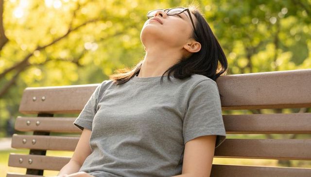 Asian woman sitting on bench tilting head back and gazing up under sunlit park canopy