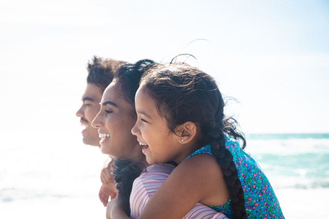 This image captures a joyful family moment at the beach, with a mother giving her daughter a piggyback ride while walking alongside the father. Perfect for use in advertisements, travel brochures, family-oriented content, and lifestyle blogs to convey themes of happiness, bonding, and outdoor fun.