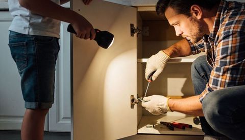 Father and child repairing kitchen cabinet hinge with flashlight and screwdriver