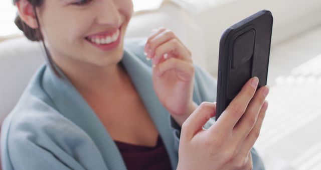 A woman in a casual outfit is holding a smartphone and smiling while engaging in a video chat. The background suggests a cozy home environment. This image is perfect for use in advertisements or blog posts about technology, communication, staying connected, or home lifestyle.