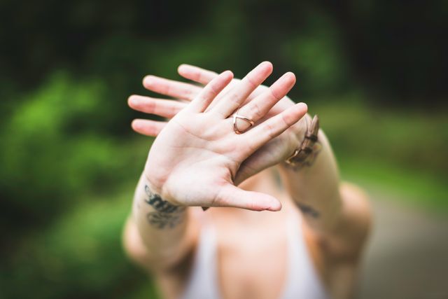 Woman in casual attire covering her face with her hands outdoors, surrounded by greenery. She is wearing rings, a wristband, and has tattoos on her arms. This can be used in themes of privacy, shyness, natural beauty, or self-expression.