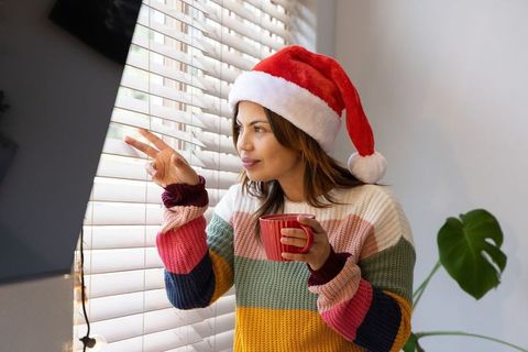 Cozy holiday morning: woman wearing santa hat peering through blinds holding red mug