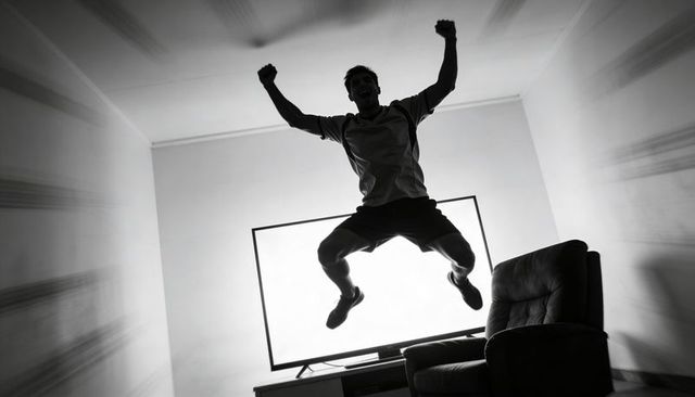 Backlit jumping man silhouette celebrating in living room with glowing tv