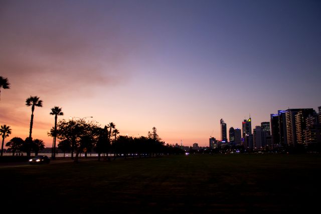 Silhouette of a city skyline at sunset with palm trees and a car driving by. The scene features a beautiful twilight sky, modern buildings with lights coming on, and an open grassy area. This image is ideal for use in travel brochures, city marketing materials, and evening outdoor events promotions. It evokes a calm, transitional time of day showcasing urban beauty and relaxation.