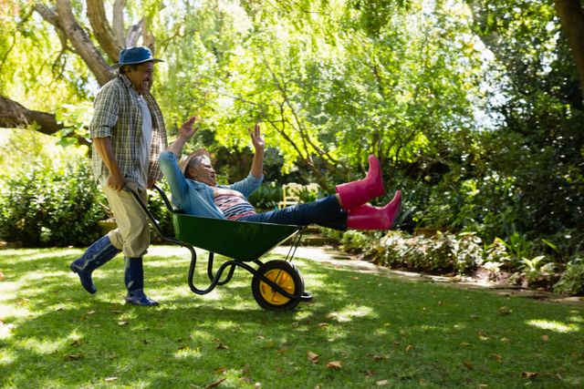Senior couple having fun in garden with man giving woman a ride in a wheelbarrow. Perfect for illustrating active retirement, outdoor leisure, and joyful moments in nature. Ideal for use in advertisements, lifestyle blogs, and retirement community promotions.