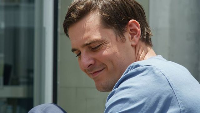 Sunlit contemplative man sitting on urban balcony wearing light blue t-shirt, looking down