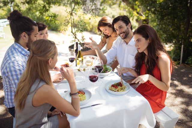 Group of friends sitting at a table outdoors, enjoying lunch and engaging in lively conversation. They are surrounded by nature, with wine glasses and plates of food in front of them. Ideal for use in advertisements for restaurants, social gatherings, or lifestyle blogs focusing on friendship and leisure activities.
