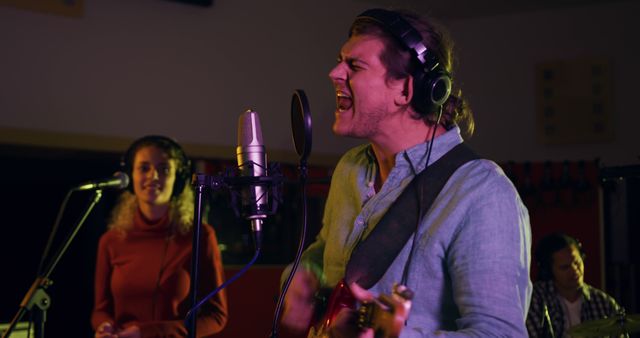 Three musicians are performing in a recording studio. The singer and guitarist is in the foreground, passionately singing into a microphone while playing the guitar. A female singer stands in the background, engaging with the performance, and another musician is visible, possibly playing drums or another instrument. This visual can be used to represent live music recording, teamwork in music production, or promotional material for music studios.