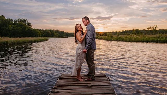 Couple embracing on wooden dock at lakeside sunset, barefoot woman in white dress