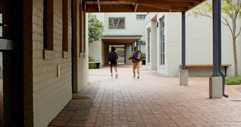 Two Male Schoolmates Walking Along Brick Corridor with Backpacks