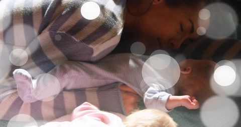 Mother resting while holding newborn on soft blanket with striped layers and sunlit bokeh