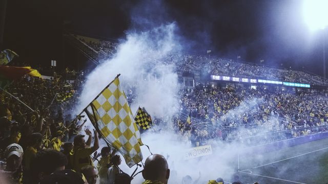 Capture of an enthusiastic crowd at a night soccer match, showing fans cheering passionately, waving flags, and smoke enhancing the atmosphere. Perfect for use in sports publications, event promotions, and advertisements highlighting the vibrant energy of live sports events.