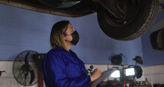 Female mechanic wearing face mask holding clipboard and inspecting the car at a car service station. automobile repair service during covid-19 pandemic