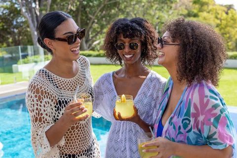 Diverse friends laughing by pool holding citrus cocktails wearing coverups and sunglasses