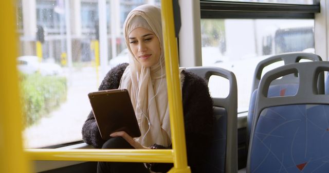 Muslim woman wearing hijab using tablet while riding public bus. Ideal for content on technology use, public transportation, modern lifestyle, and cultural representation. Perfect for travel blogs, technology websites, and marketing materials highlighting digital device use in everyday life.