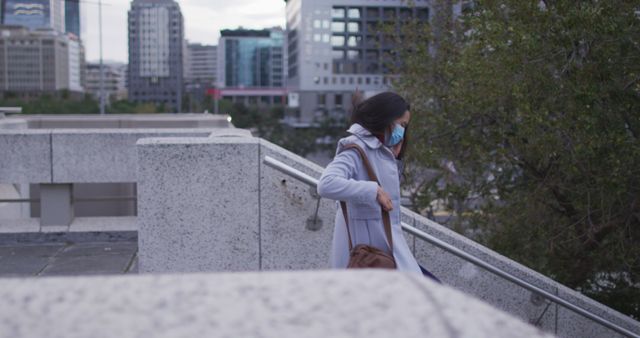 Woman in city wearing face mask walking down stairs. Urban background with buildings and greenery. Ideal for conveying topics related to urban life, daily commute, pandemic precautions, public health, and safety measures.