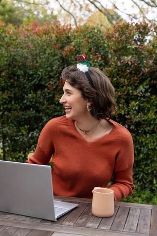 Young woman laughing while working outdoors with festive headband, laptop and mug
