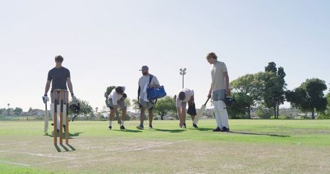 Cricket team preparing pitch: players marking crease with bats helmets pads on sunny field
