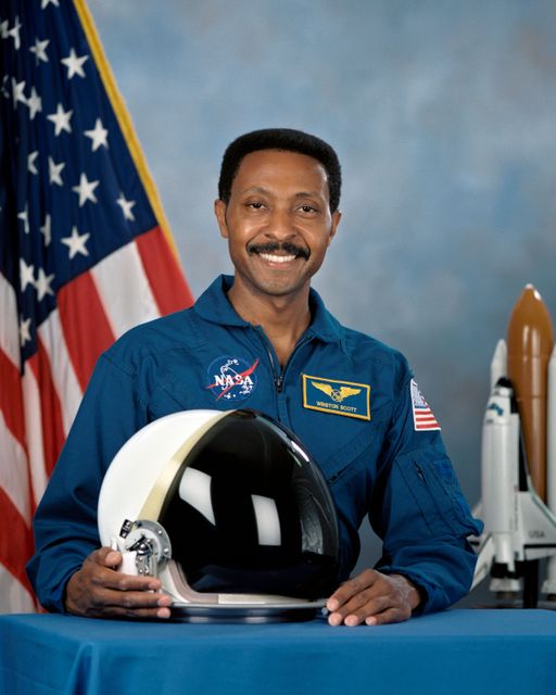 Image shows an astronaut smiling in a NASA space suit and holding a helmet with an American flag in the background. This image is ideal for use in educational materials, space exploration articles, and promoting diversity in STEM fields.