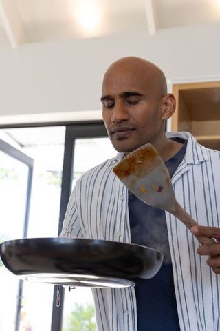 Man cooking in sunlit kitchen smelling spatula with steaming nonstick pan morning routine