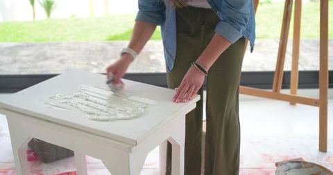 Mature woman applying white primer to wooden side table in bright home studio