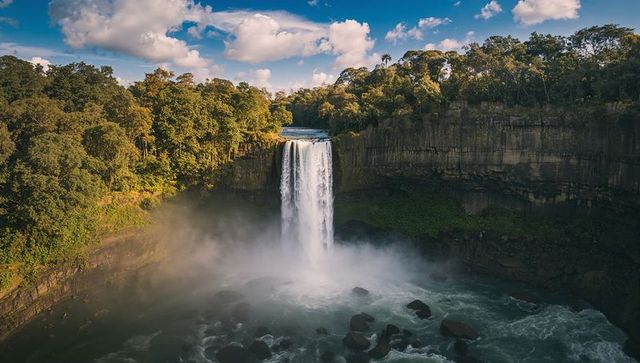 Plunging single-drop waterfall cascading into misty river gorge surrounded by lush forest