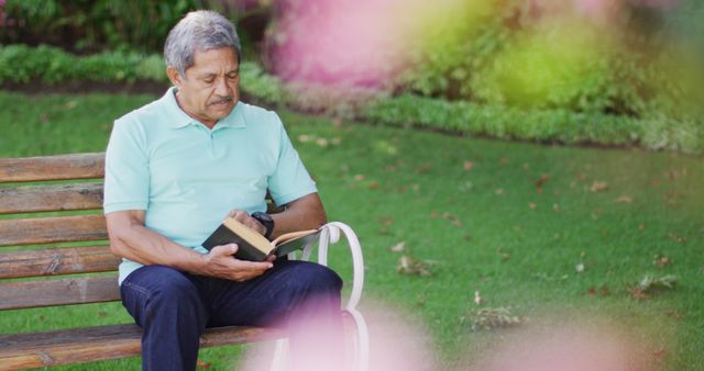 Mature man sitting on bench reading book, surrounded by green lawn in outdoor park setting. Ideal for use in advertisements for senior leisure products, relaxation tips, promoting healthy lifestyle, or content focused on enjoying nature.