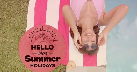 Smiling woman sunbathing poolside in pink swimsuit on striped towel with sunglasses
