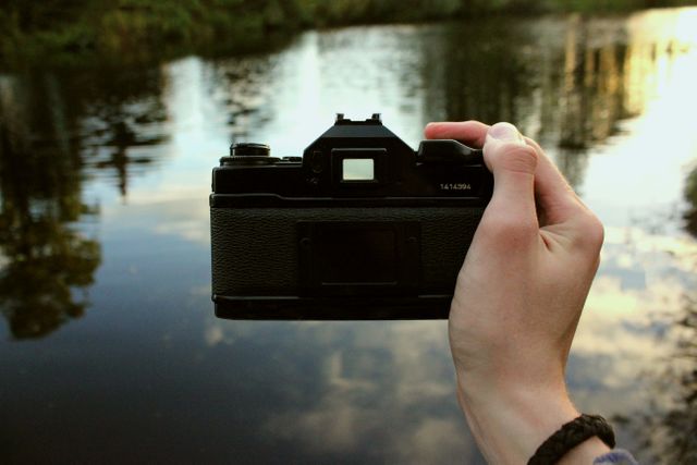 Hand holding a camera by calm lake during sunset with water reflecting surroundings. Perfect for themes of photography, nature, creativity, and outdoor activities. Ideal for blogs, articles on hobbies, creativity inspiration, and promotional material for photography equipment.