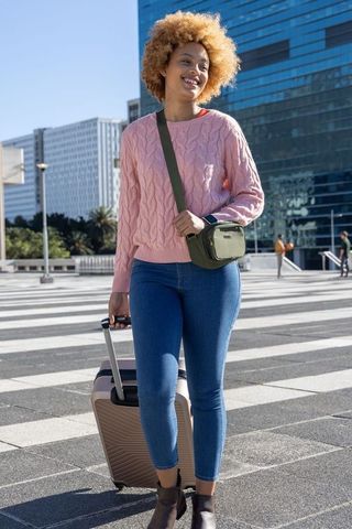 African american woman walking with roller suitcase in urban plaza wearing pink sweater