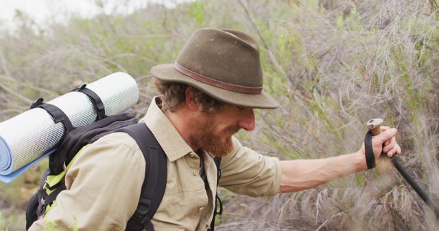 Image shows a bearded man hiking through a wilderness area, wearing a green hat and carrying a backpack with a rolled mat. He appears focused, using a trekking pole while navigating the outdoor terrain. Ideal for anything related to outdoor adventures, trekking gear, nature exploration, or lifestyle blogs focused on travel and camping.