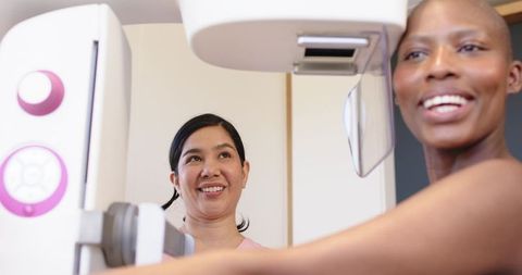 Smiling hispanic patient undergoing mammogram while african american technician adjusts equipment in