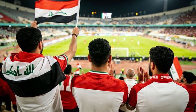 Iraqi soccer fans waving national flag in stadium at night cheering in stands