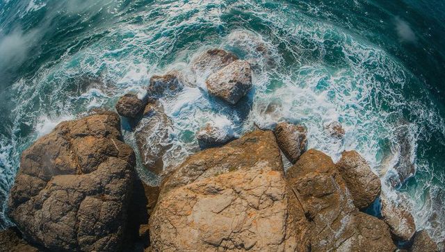 Aerial view of rugged rocky coastline with teal sea crashing into tan boulders and white surf