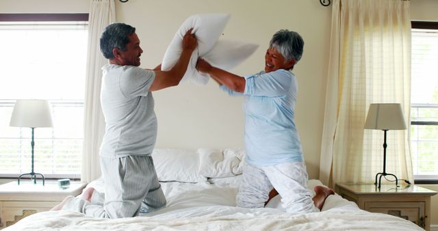 A middle-aged Asian couple is playfully engaging in a pillow fight on a bed, with copy space. Their joyful activity suggests a lighthearted and loving relationship, capturing a moment of fun and intimacy.