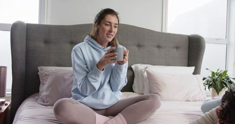 Woman sitting cross-legged on bed holding mug in cozy pastel bedroom relaxing morning