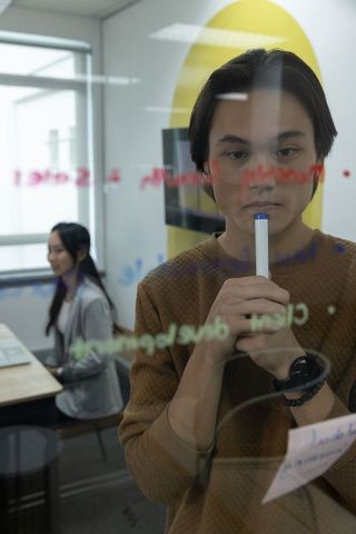 Young professional brainstorming on glass wall with marker while coworker using laptop