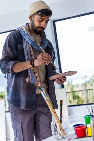 Indian male artist painting in bright studio wearing beanie and scarf with palette