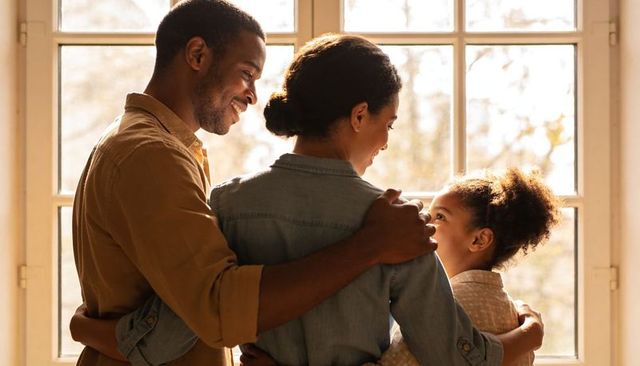Parents embracing daughter at sunlit window, warm family hug in cozy home interior