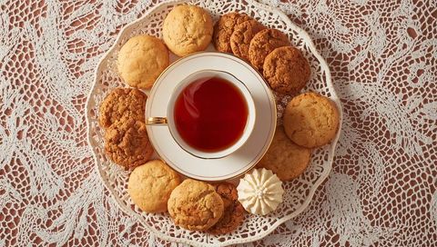 Vintage porcelain teacup with gold rim encircled by assorted cookies on lace tablecloth