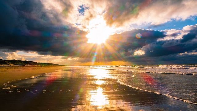 Golden sunburst over coastal beach reflecting on wet sand with dramatic clouds and sunbeams
