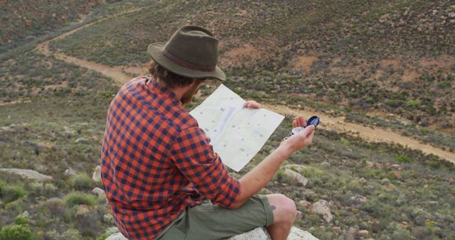 Caucasian male survivalist sitting on mountain peak in wilderness, holding map and compass. exploration, travel and adventure, survivalist in nature.