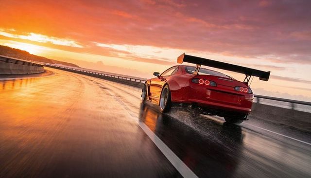 Red sports coupe sprinting along wet coastal highway at sunset with giant rear wing