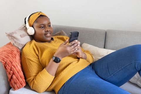 African american teen reclining on sofa listening with headphones and using flip phone